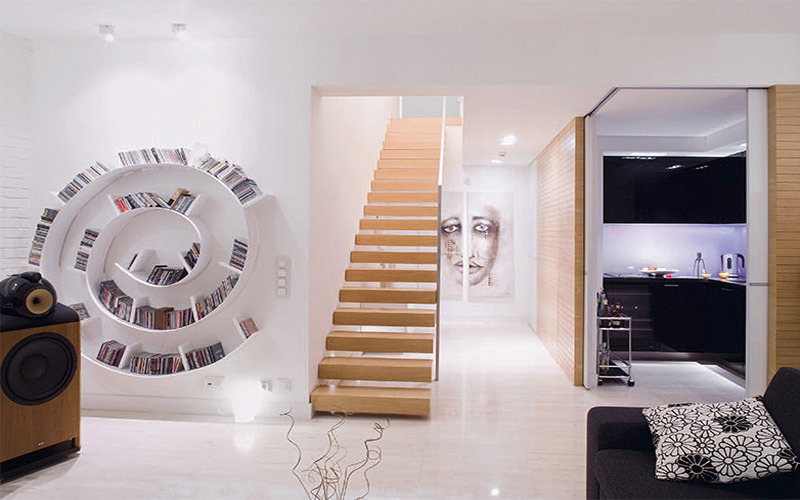 Białe wnętrze domu z spiralną biblioteczką na płyty CD, drewnianymi schodami i widokiem na czarną kuchnię z podświetleniem LED