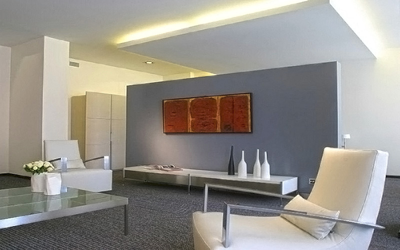 Nowoczesne wnętrze biurowe z minimalistycznym wystrojem, szarym panelem ściennym, abstrakcyjnym obrazem, białymi fotelami i szklanym stolikiem kawowym.