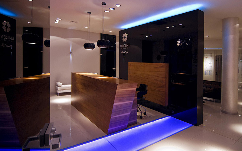 Nowoczesne wnętrze recepcji z drewnianymi ladami, podświetleniem LED w kolorze niebieskim, czarnymi panelami ściennymi z logo firmy Unident Union i designerskimi lampami wiszącymi.