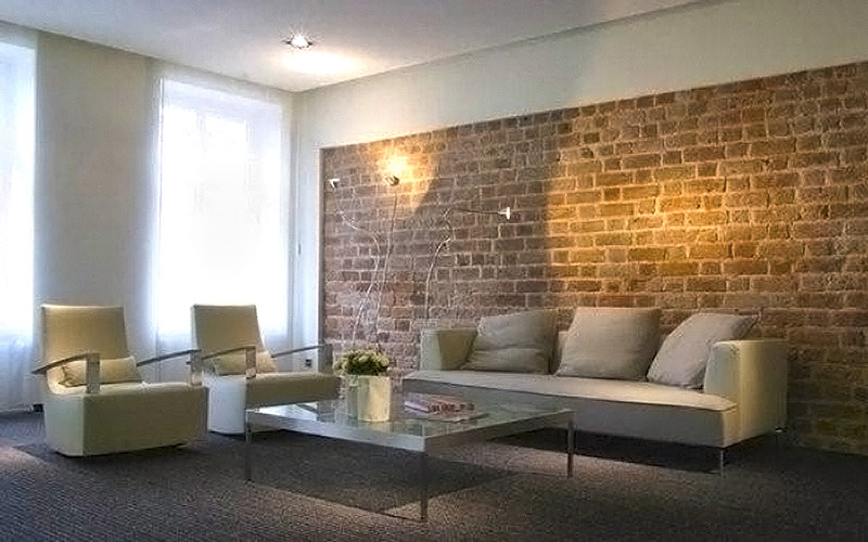 Nowoczesny salon z ceglaną ścianą, jasną sofą i fotelami, szklanym stolikiem kawowym oraz dekoracyjnymi lampami ściennymi.