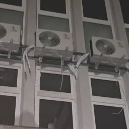 EKO- KLIMA - Trzy zewnętrzne jednostki klimatyzacyjne zamontowane na elewacji budynku z widocznymi przewodami i wspornikami mocującymi.