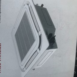 EKO- KLIMA - Klimatyzator kasetonowy sufitowy, widok z boku, z widoczną kratką wentylacyjną i obudową w kolorze białym, na tle białej ściany.