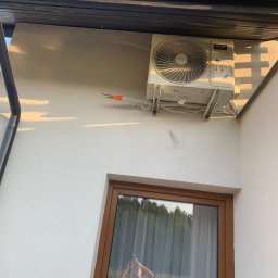 EKO- KLIMA - Biała jednostka zewnętrzna klimatyzacji AUX zamontowana na ścianie budynku, widoczne przewody i odbicie osoby robiącej zdjęcie w oknie.