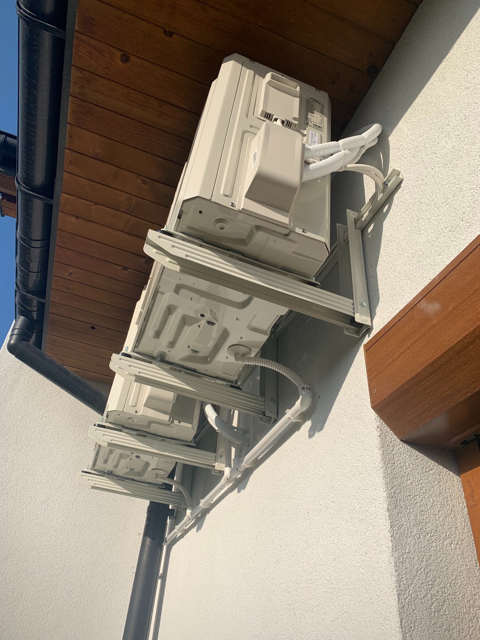 Zewnętrzna jednostka klimatyzacji zamontowana na ścianie budynku, widok z dołu, z widocznymi przewodami i elementami montażowymi.