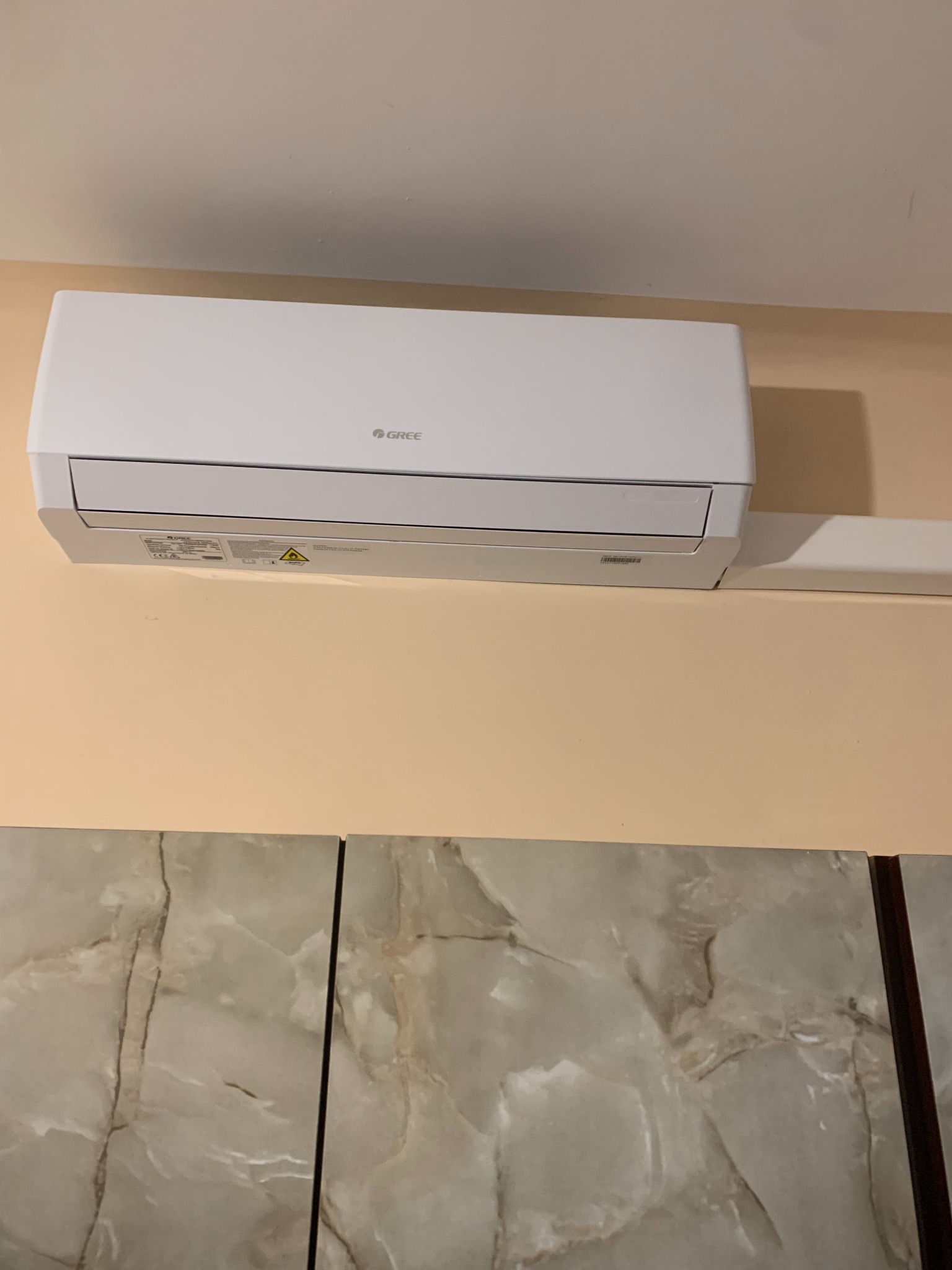Biała jednostka wewnętrzna klimatyzacji marki Gree zamontowana na beżowej ścianie, widoczne płytki imitujące marmur na dolnej części kadru.