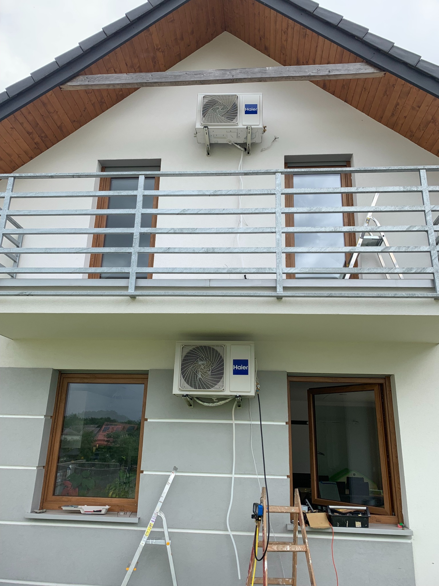 Dwa klimatyzatory Haier zamontowane na elewacji dwukondygnacyjnego domu z balkonem, drabiny i narzędzia sugerują montaż lub serwis.