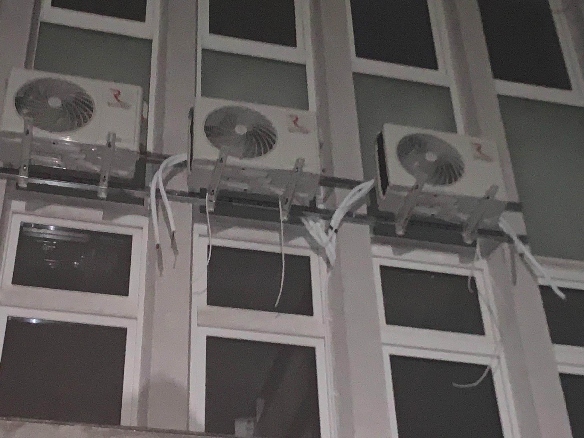 Trzy zewnętrzne jednostki klimatyzacyjne zamontowane na elewacji budynku z widocznymi przewodami i wspornikami mocującymi.