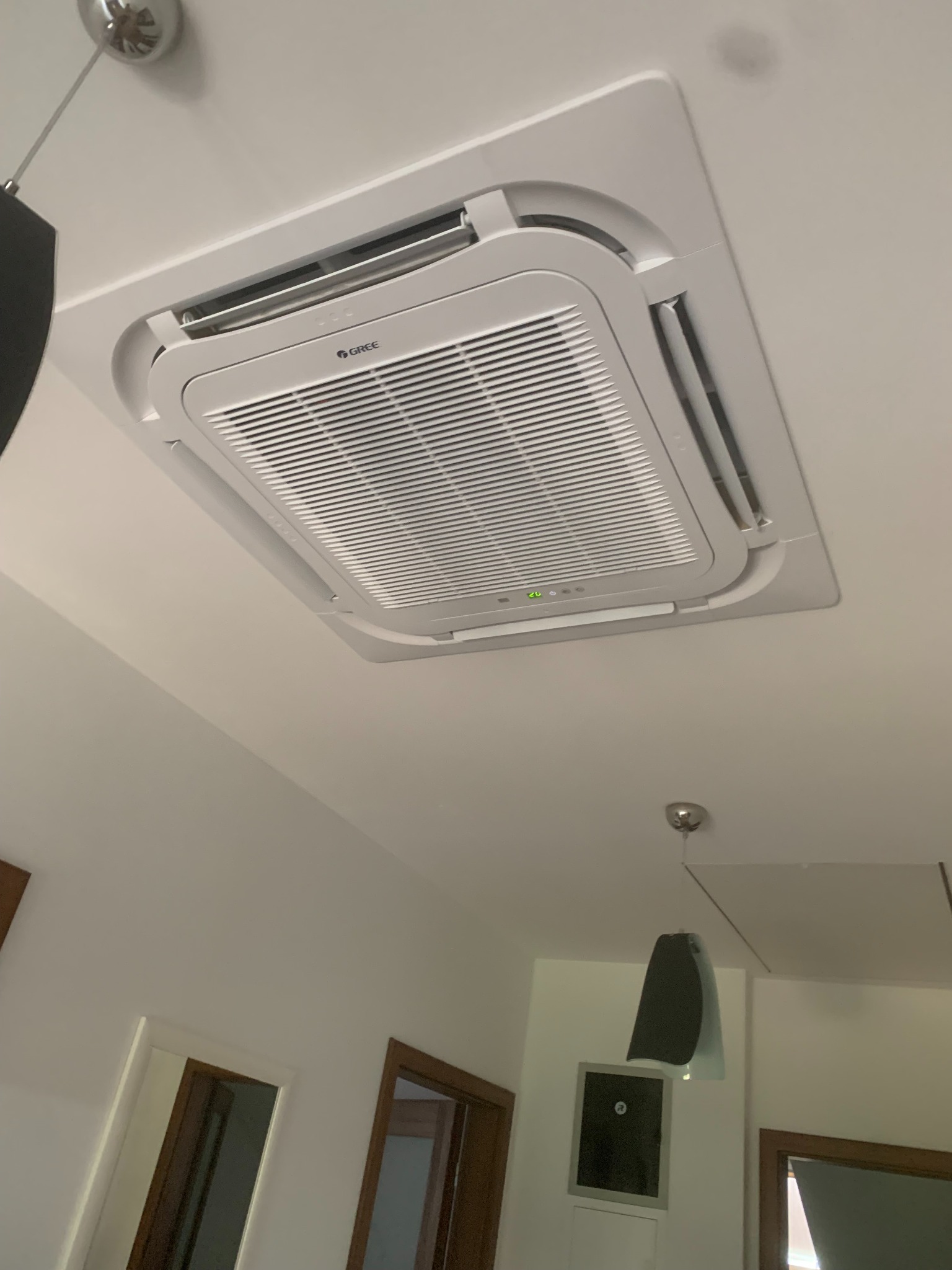 Kasetonowa jednostka klimatyzacyjna marki Gree zamontowana w suficie podwieszanym w pomieszczeniu z widocznymi drzwiami i lampą.