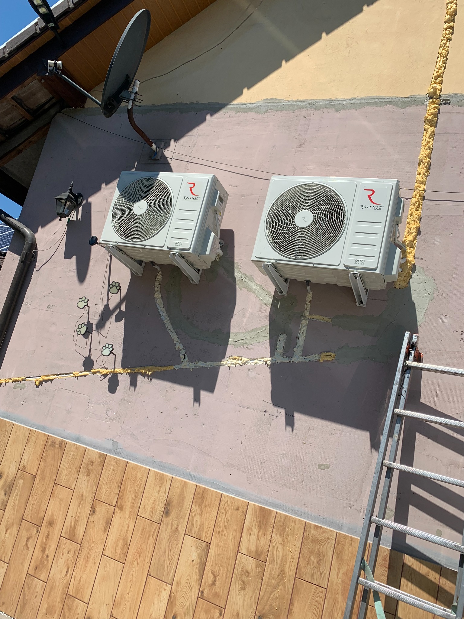 Dwa klimatyzatory Rotenso zamontowane na elewacji budynku, z widoczną pianką montażową i drabiną. Ślady po instalacji. Ujęcie z dołu, z ukośną perspektywą.
