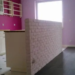 Kuchnia z wyspą oddzieloną ścianką z białej cegły, widoczne białe szafki, fioletowa ściana i fragment nowej podłogi.