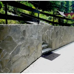 Kamienny murek oporowy z nieregularnych płyt, schodki prowadzące w górę, czarna wycieraczka. Tło: zieleń drzew i fragment dachu budynku.