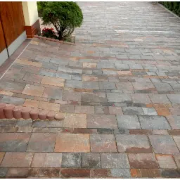 Ułożona kostka brukowa w odcieniach szarości i brązu tworząca podjazd z obrzeżami. Widoczna zieleń roślinności przy budynku.