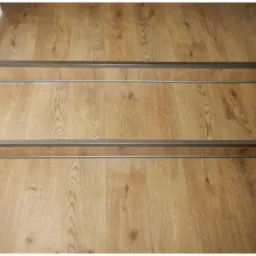 Drewnopodobne schody z metalowymi listwami antypoślizgowymi, widok z góry. Jasne drewno z wyraźnym usłojeniem. Minimalistyczne wykończenie.