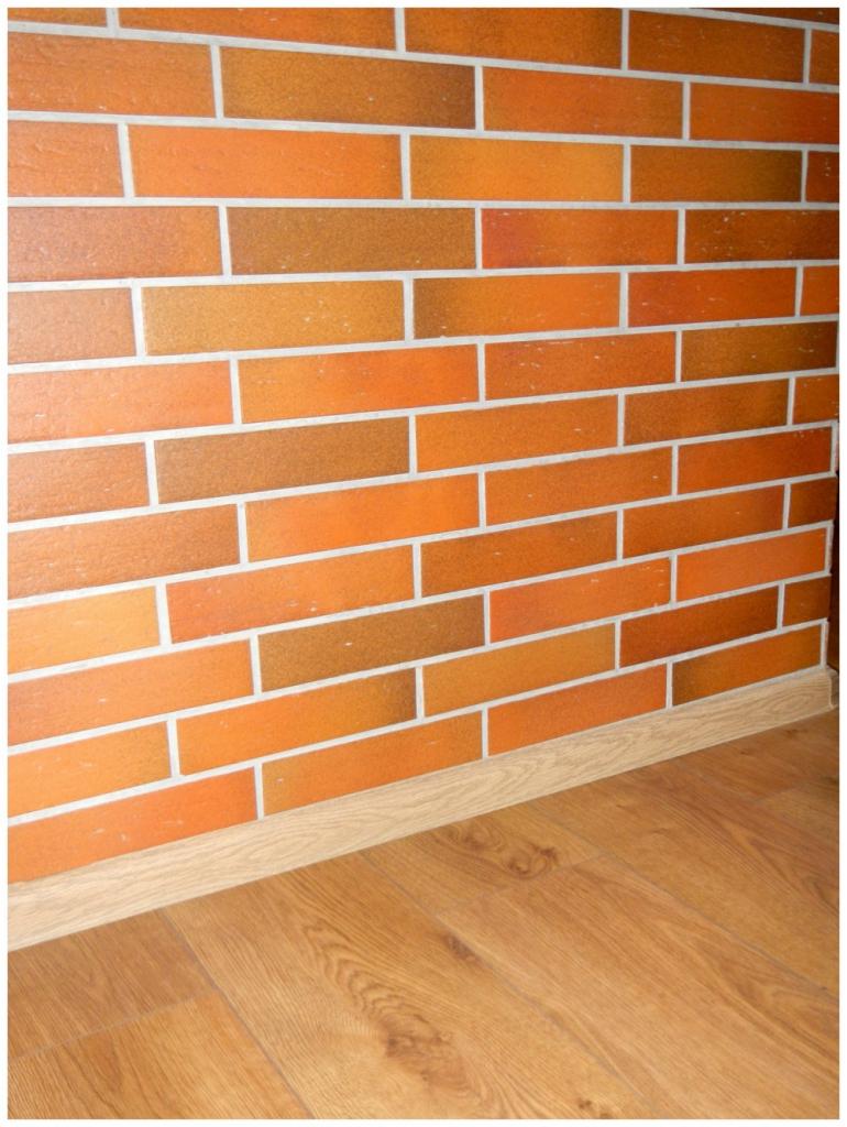 Ściana z dekoracyjnej cegły w odcieniach pomarańczy i brązu, z jasną fugą i drewnianą podłogą. Widoczna listwa przypodłogowa.