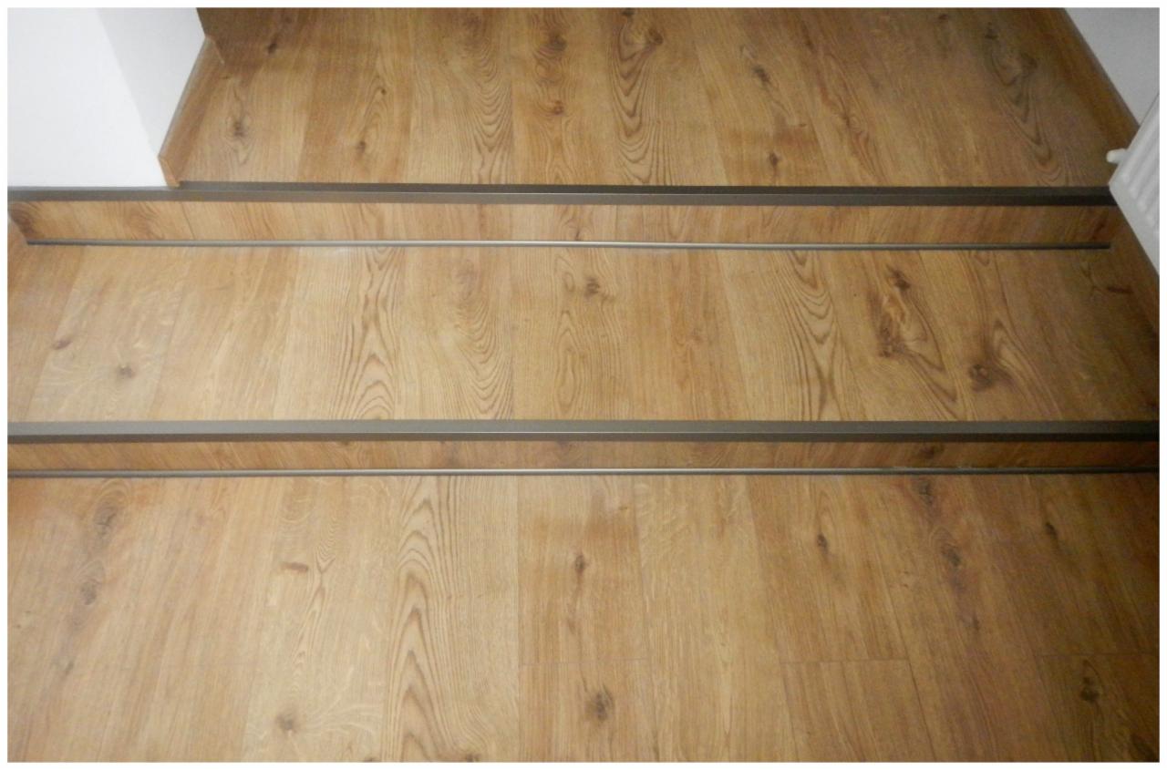 Drewnopodobne schody z metalowymi listwami antypoślizgowymi, widok z góry. Jasne drewno z wyraźnym usłojeniem. Minimalistyczne wykończenie.