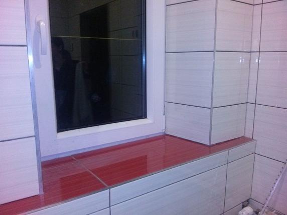 Wykończenie wnęki okiennej w łazience z użyciem czerwonych płytek ceramicznych, ściany wyłożone jasnymi płytkami imitującymi drewno z ciemnymi fugami.