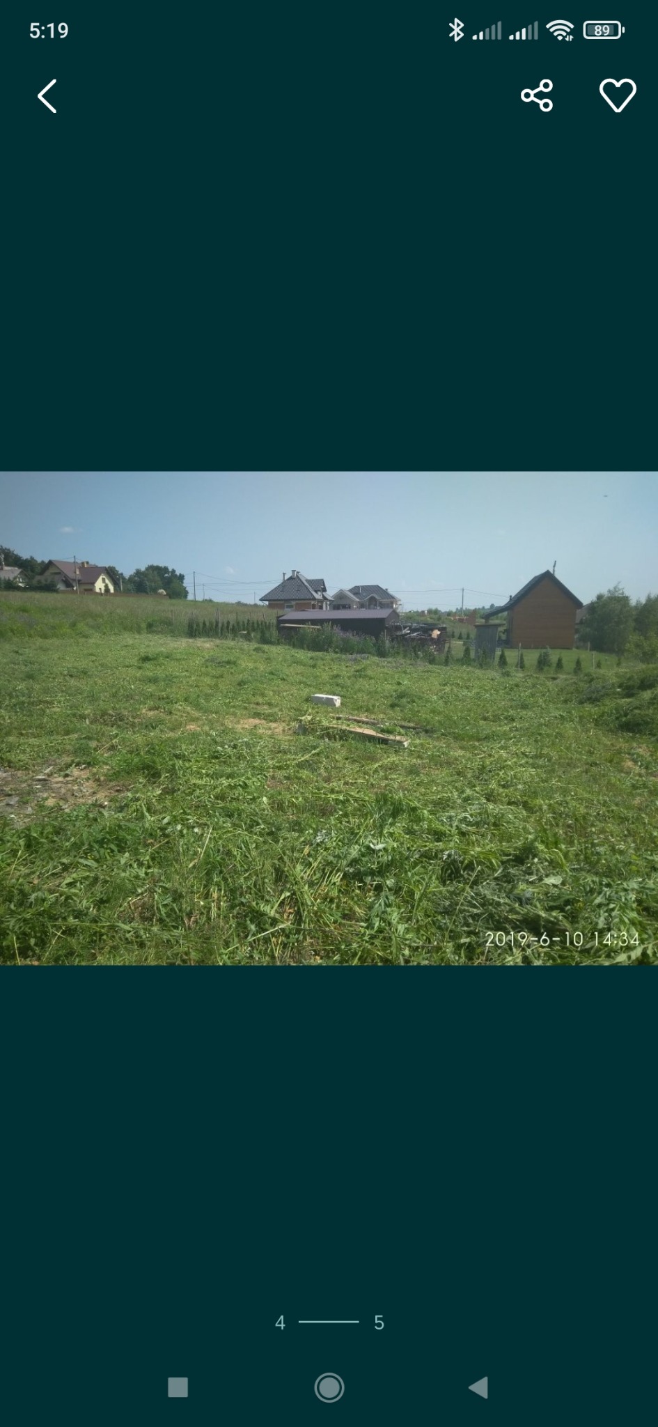 Nierównomiernie skoszona trawa na dużej działce z widocznymi budynkami mieszkalnymi w tle, wykonane w słoneczny dzień.