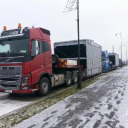 Czerwona ciężarówka Volvo FH16 z naczepą przewożąca duży kontener na niskopodwoziowej platformie, zaparkowana na poboczu drogi w zimowy dzień, z widocznymi innymi ciężarówkami w tle.
