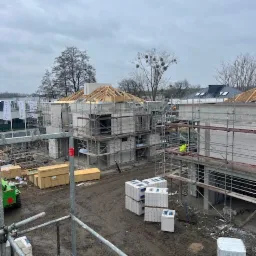 Wznoszenie budynków z betonu komórkowego na placu budowy, widoczne rusztowania, drewniane elementy konstrukcji dachu i palety z materiałami budowlanymi na błotnistym terenie.