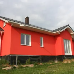 Czerwony dom jednorodzinny po remoncie elewacji z białymi oknami i ciemną dachówką, widok z boku na tle pochmurnego nieba i zieleni.