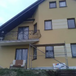 Ocieplona elewacja domu dwukondygnacyjnego w odcieniach beżu, z dekoracyjnymi pasami. Widoczne rusztowania i zabezpieczone okna folią malarską.