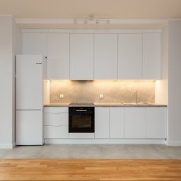 Grast & MTB Designer Sp. z o.o. - Nowoczesna, jasna kuchnia z białymi szafkami, szarym blatem i podświetleniem LED, widok na balkon przez duże okno.