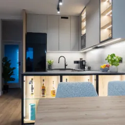 Nowoczesna kuchnia w Warszawie z szarymi szafkami, czarną armaturą i oświetleniem LED, z wyspą z podświetlanymi półkami, na których stoją butelki alkoholu. Na blacie doniczka z ziołami i misa...