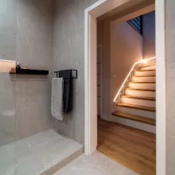 Nowoczesna łazienka z szarymi płytkami imitującymi marmur, czarną półką z kosmetykami, wieszakiem na ręczniki i widokiem na schody z podświetleniem LED przez białe drzwi.