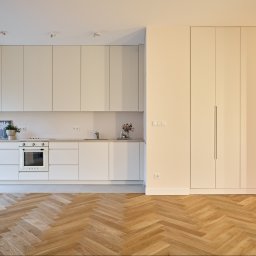 Grast & MTB Designer Sp. z o.o. - Jasna kuchnia z parkietem w jodełkę. Minimalistyczne białe szafki, sprzęt AGD SMEG, przeszklone drzwi balkonowe. Nowoczesne wykończenie wnętrza.