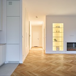 Grast & MTB Designer Sp. z o.o. - Jasne wnętrze z parkietem w jodełkę: biała zabudowa kuchenna, wnęki z oświetleniem i dekoracjami, imitacja kominka. Minimalistyczny design, ciepłe światło.