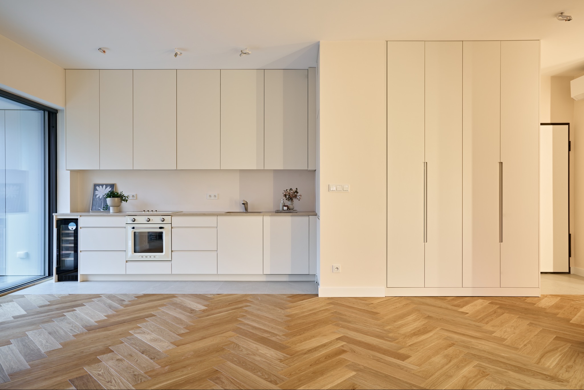 Jasna kuchnia z parkietem w jodełkę. Minimalistyczne białe szafki, sprzęt AGD SMEG, przeszklone drzwi balkonowe. Nowoczesne wykończenie wnętrza.