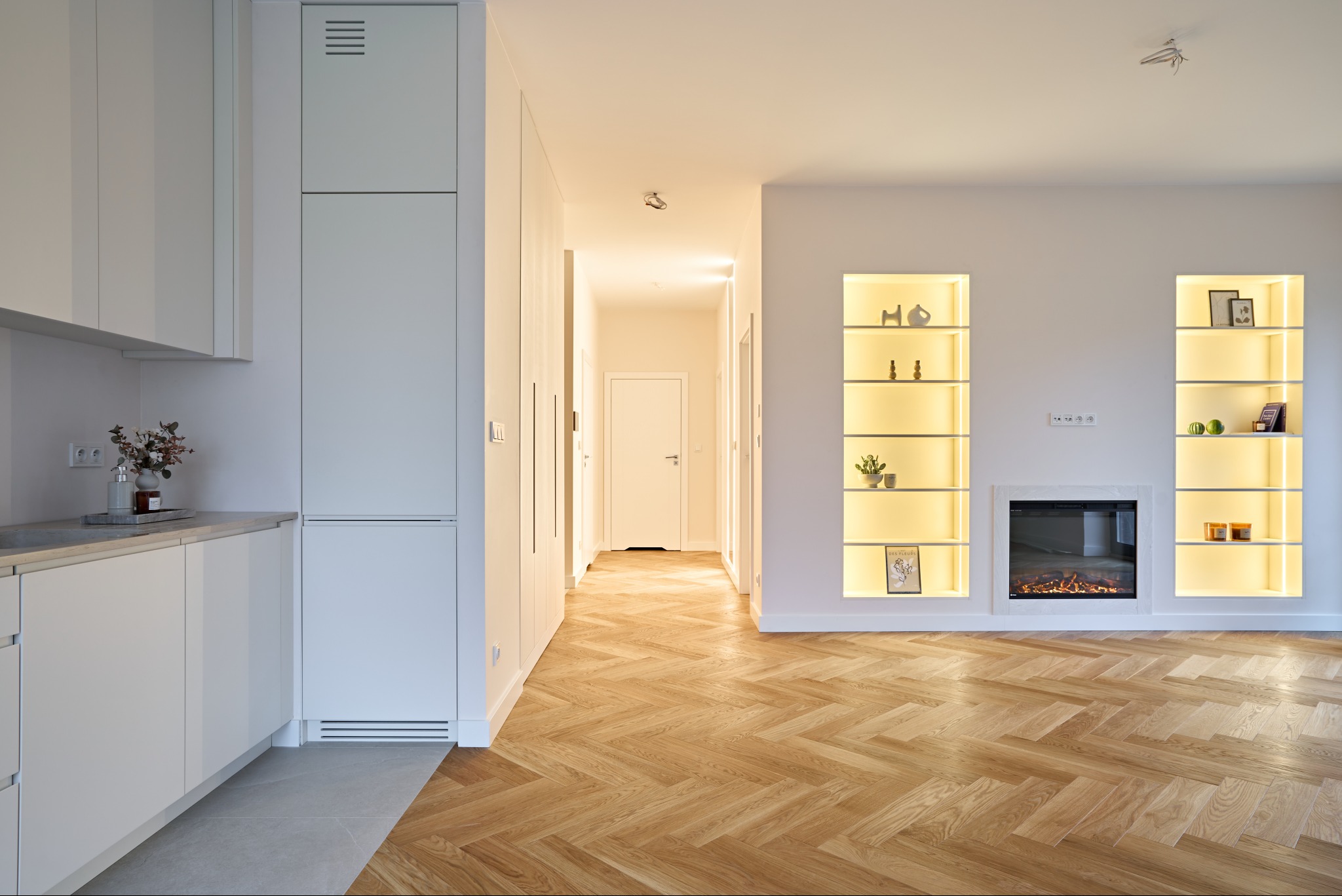 Jasne wnętrze z parkietem w jodełkę: biała zabudowa kuchenna, wnęki z oświetleniem i dekoracjami, imitacja kominka. Minimalistyczny design, ciepłe światło.