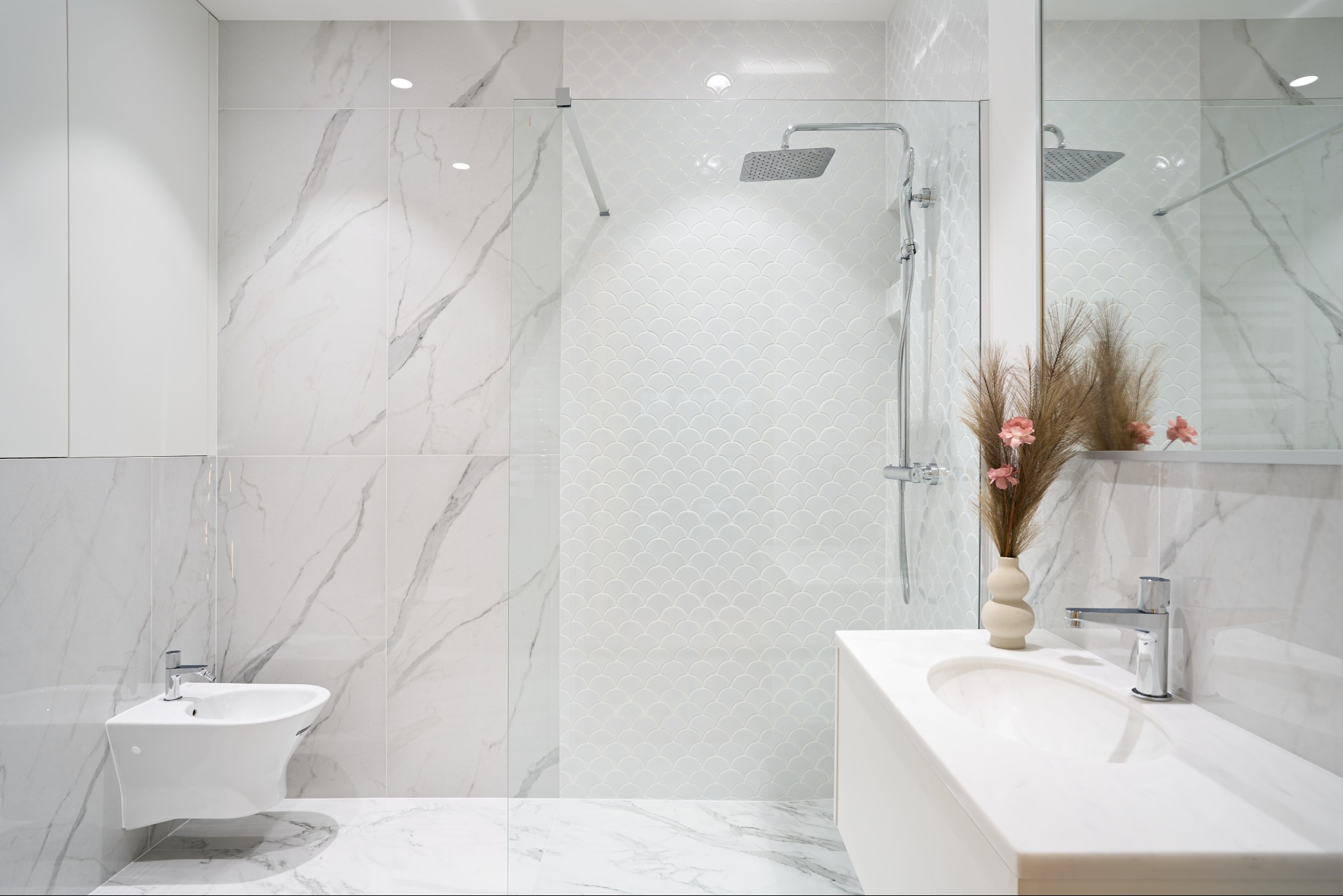 Elegancka łazienka w bieli: marmurowe płytki, kabina prysznicowa ze szklaną ścianką, umywalka z minimalistyczną armaturą i dekoracyjny wazon z kwiatami.