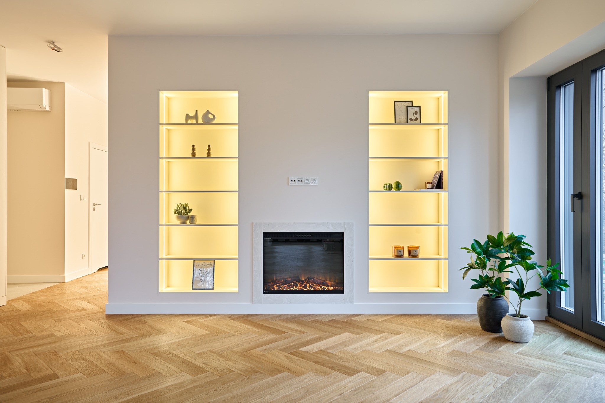 Salon z parkietem w jodełkę, oświetlonymi wnękami z dekoracjami i elektrycznym kominkiem. Minimalistyczny design z roślinami doniczkowymi przy oknie.