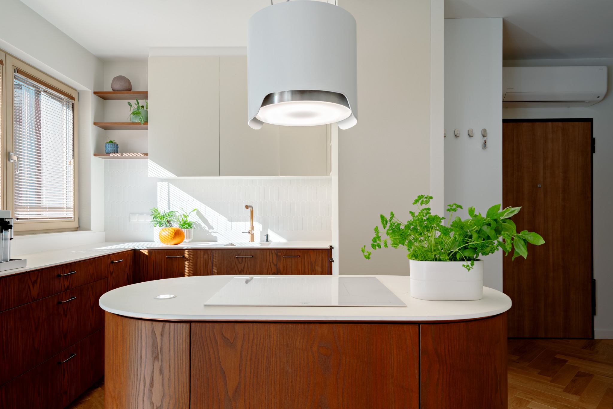 Nowoczesna kuchnia z drewnianymi szafkami, białym blatem i okrągłą wyspą. Nad wyspą designerski okap. Na blacie zioła w doniczce. Parkiet w jodełkę.