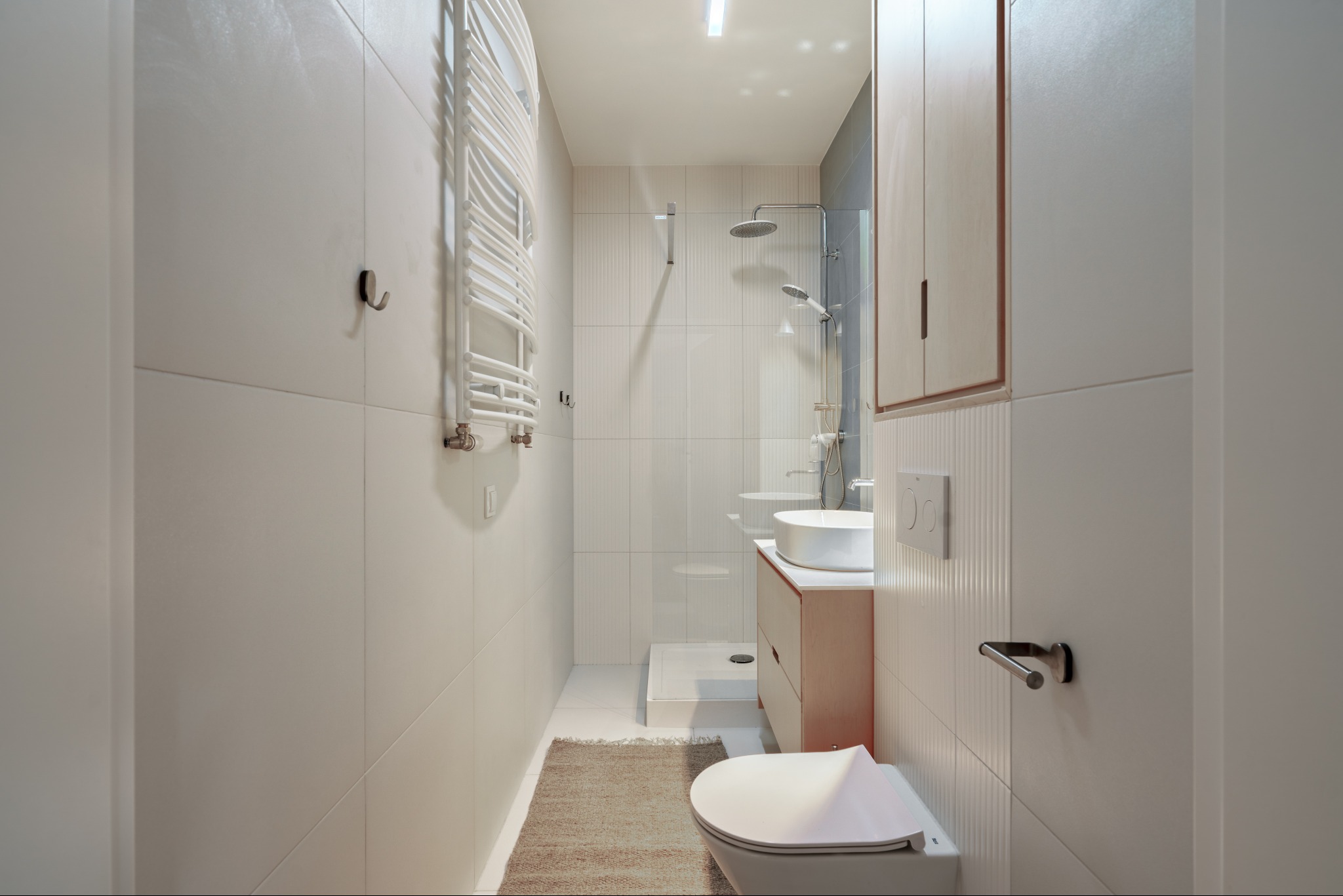 Jasna łazienka z kabiną prysznicową typu walk-in, umywalką nablatową i grzejnikiem drabinkowym. Minimalistyczny design z białymi płytkami i drewnianymi akcentami.