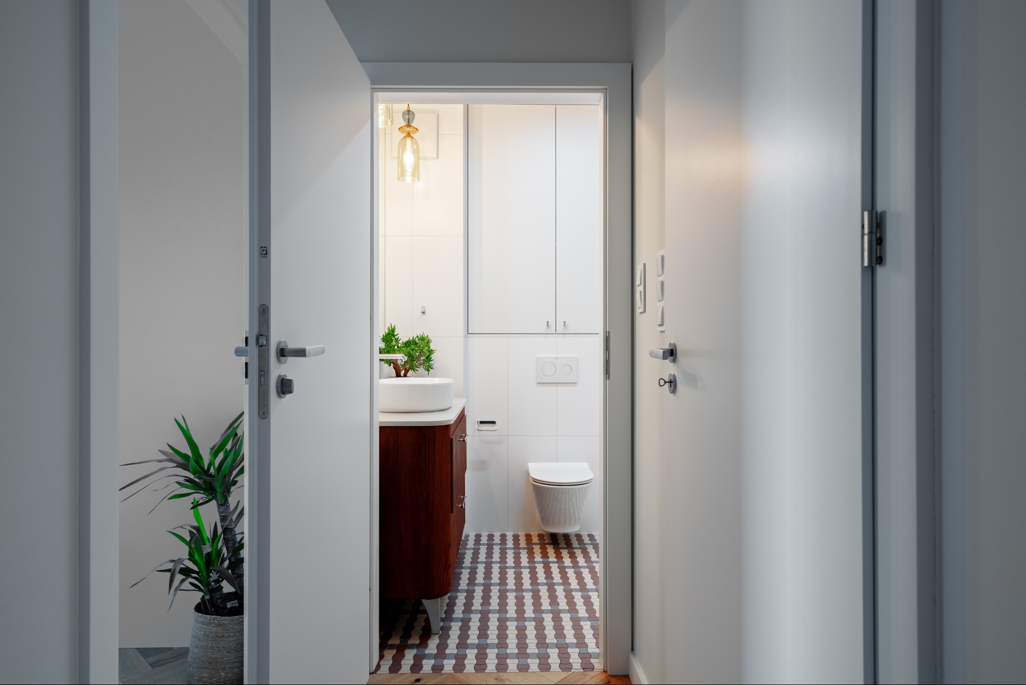 Nowoczesna łazienka z białą ceramiką, drewnianą szafką pod umywalką i wzorzystą podłogą, widziana z korytarza przez otwarte drzwi. Roślina w doniczce po lewej.