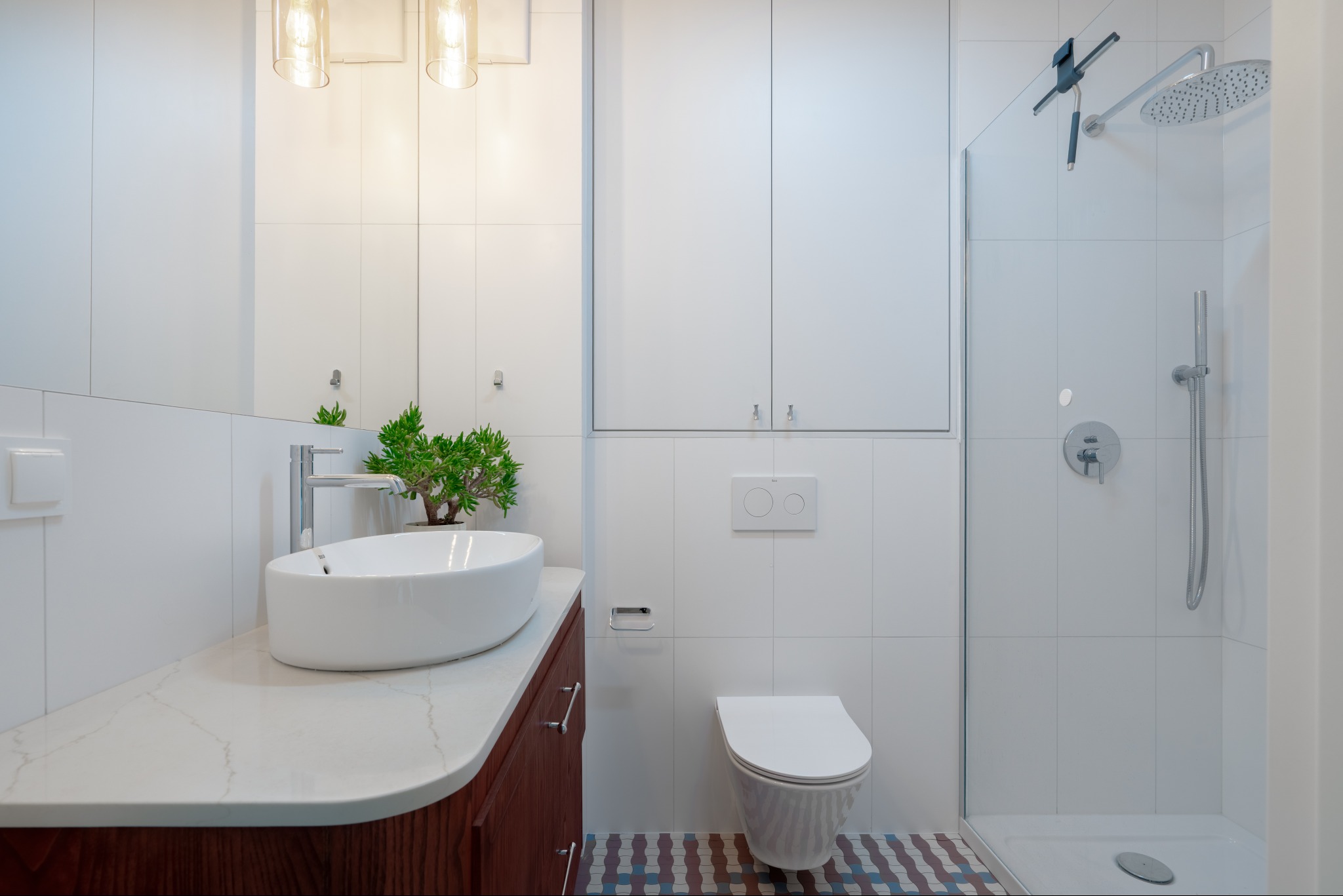 Nowoczesna łazienka z owalną umywalką na drewnianej szafce, białą toaletą i kabiną prysznicową. Uwagę przykuwają kolorowe płytki podłogowe i minimalistyczne oświetlenie.