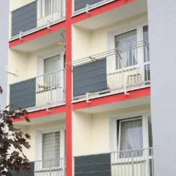 Fasada budynku z balkonami zabezpieczonymi metalowymi barierkami i szarymi panelami, widok z dołu na kilka pięter.