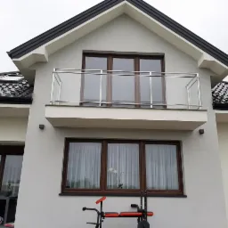 Elewacja domu z balkonem zabezpieczonym balustradą szklaną z elementami stali nierdzewnej. Widok z dołu na balkon i okna z brązowymi ramami.