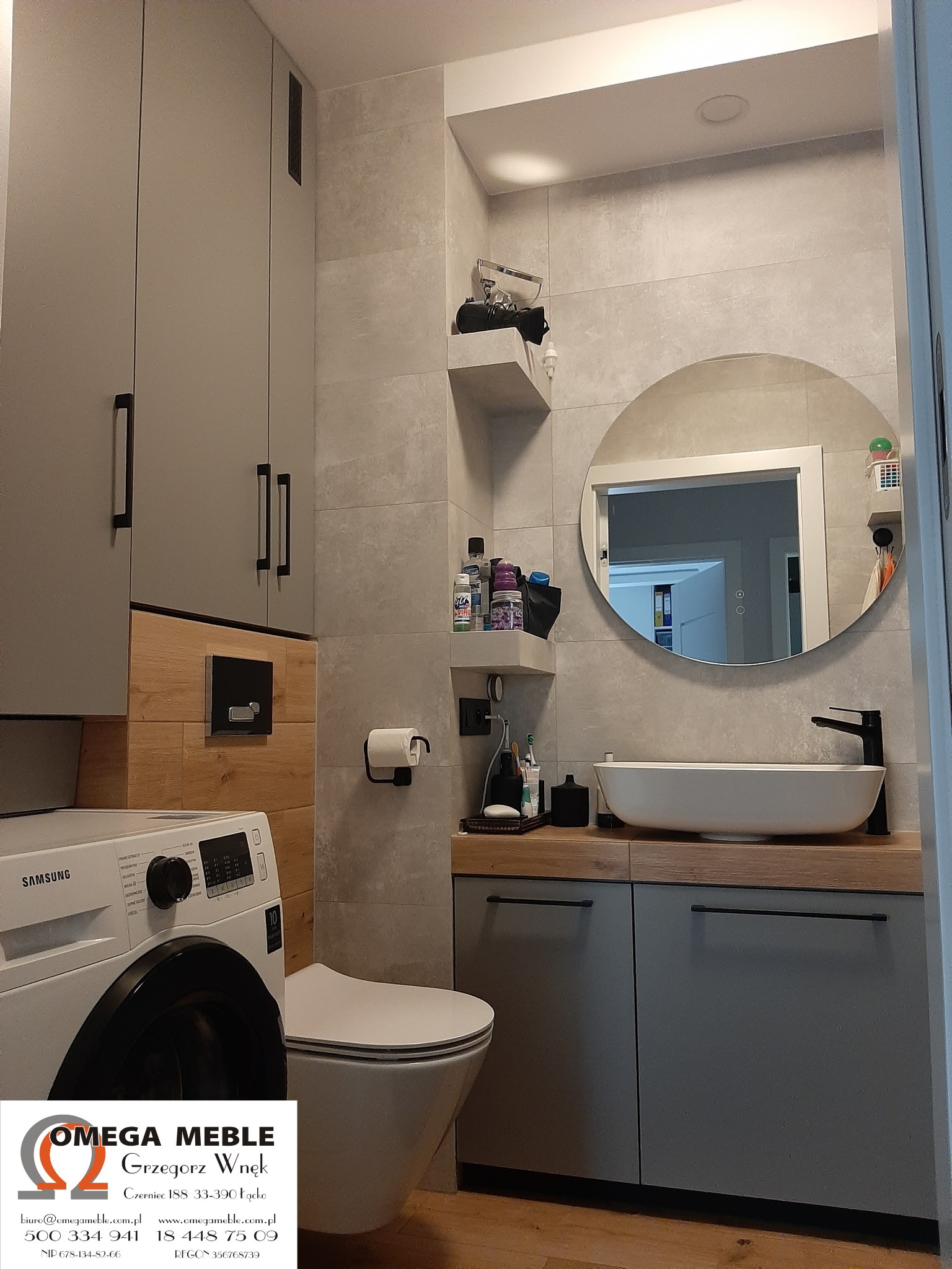 Szara łazienka z zabudową meblową pod umywalką, okrągłym lustrem, toaletą i pralką Samsung, widoczne logo firmy Omega Meble