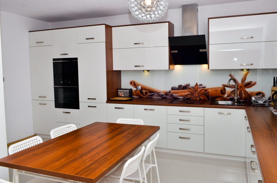 Nowoczesna kuchnia z białymi frontami i drewnianymi akcentami, oświetleniem punktowym pod szafkami górnymi i blatem z motywem kawy i cynamonu, uzupełniona drewnianym stołem i białymi krzesłami.