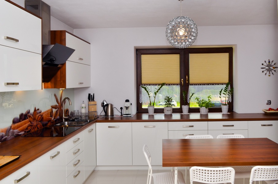 Jasna kuchnia z białymi szafkami i drewnianymi blatami, okno z roletami i roślinami, designerska lampa sufitowa.