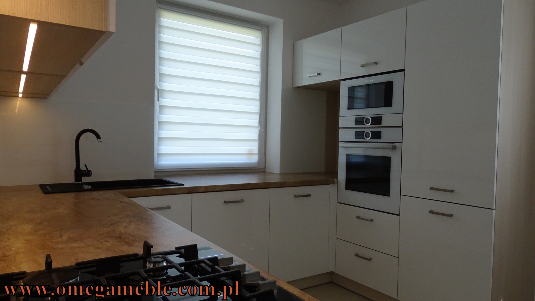 Nowoczesna kuchnia z białymi, błyszczącymi frontami, drewnianym blatem i czarną armaturą, oświetlona naturalnym światłem z okna z żaluzjami dzień-noc.