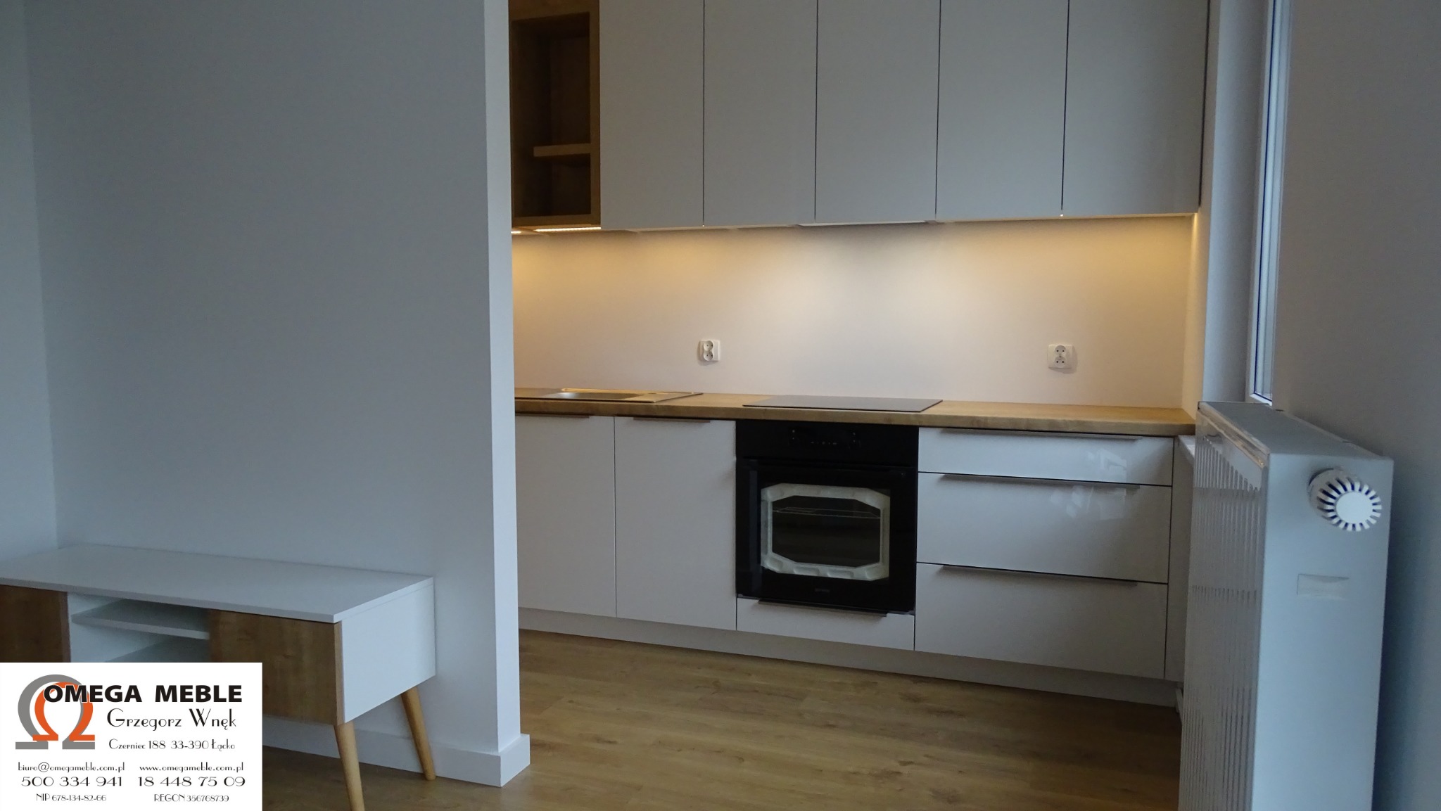 Nowoczesna kuchnia z białymi szafkami, drewnianym blatem i czarnym piekarnikiem, oświetlenie podszafkowe, widoczny fragment szafki RTV z logo firmy Omega Meble.