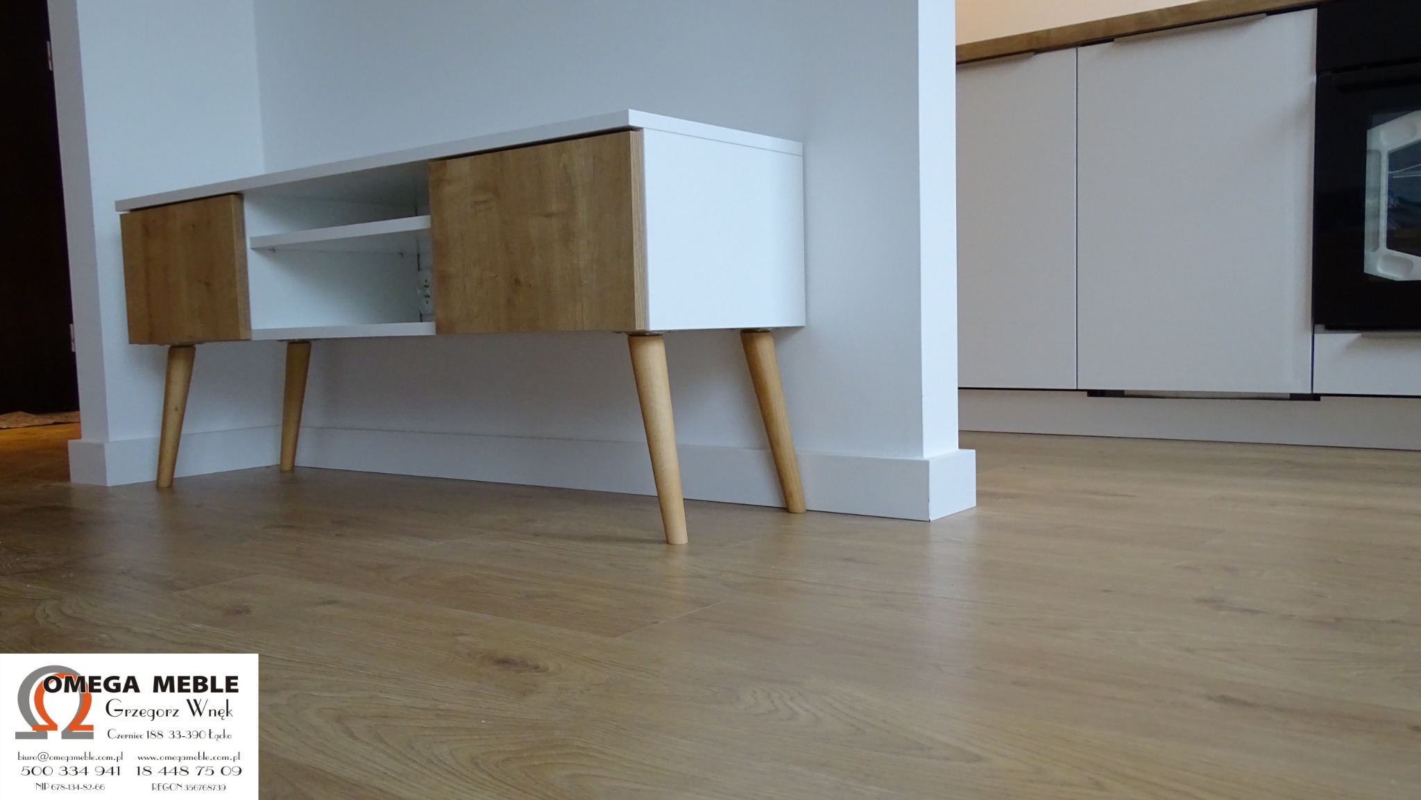 Biało-drewniana szafka RTV z nóżkami w stylu retro, umieszczona pod ścianą w nowoczesnym wnętrzu, widok z niskiej perspektywy na parkiet.