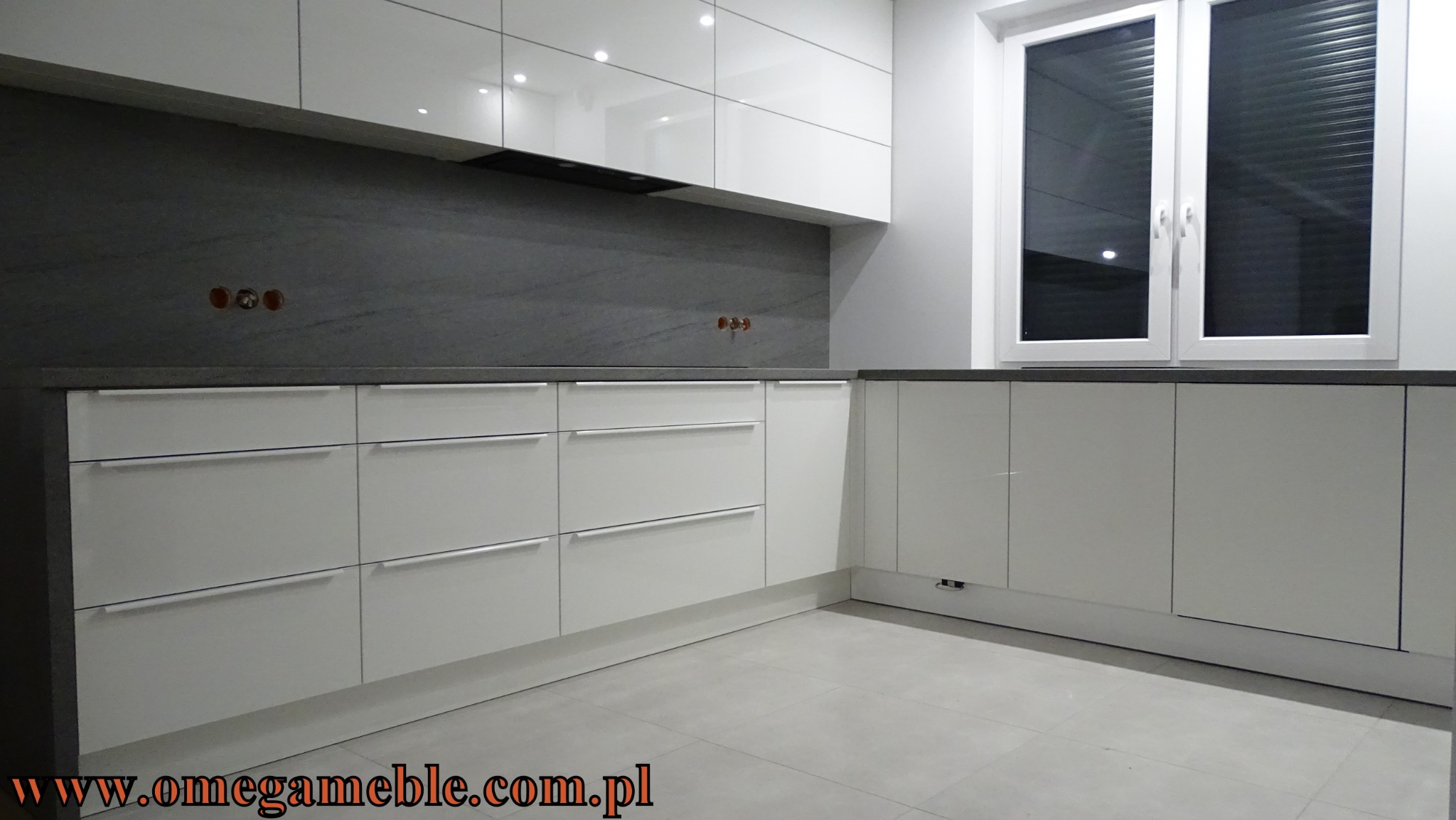 Nowoczesna, biała kuchnia z szarym blatem i backsplashiem z imitacji kamienia, widoczne gniazdka elektryczne na ścianie.