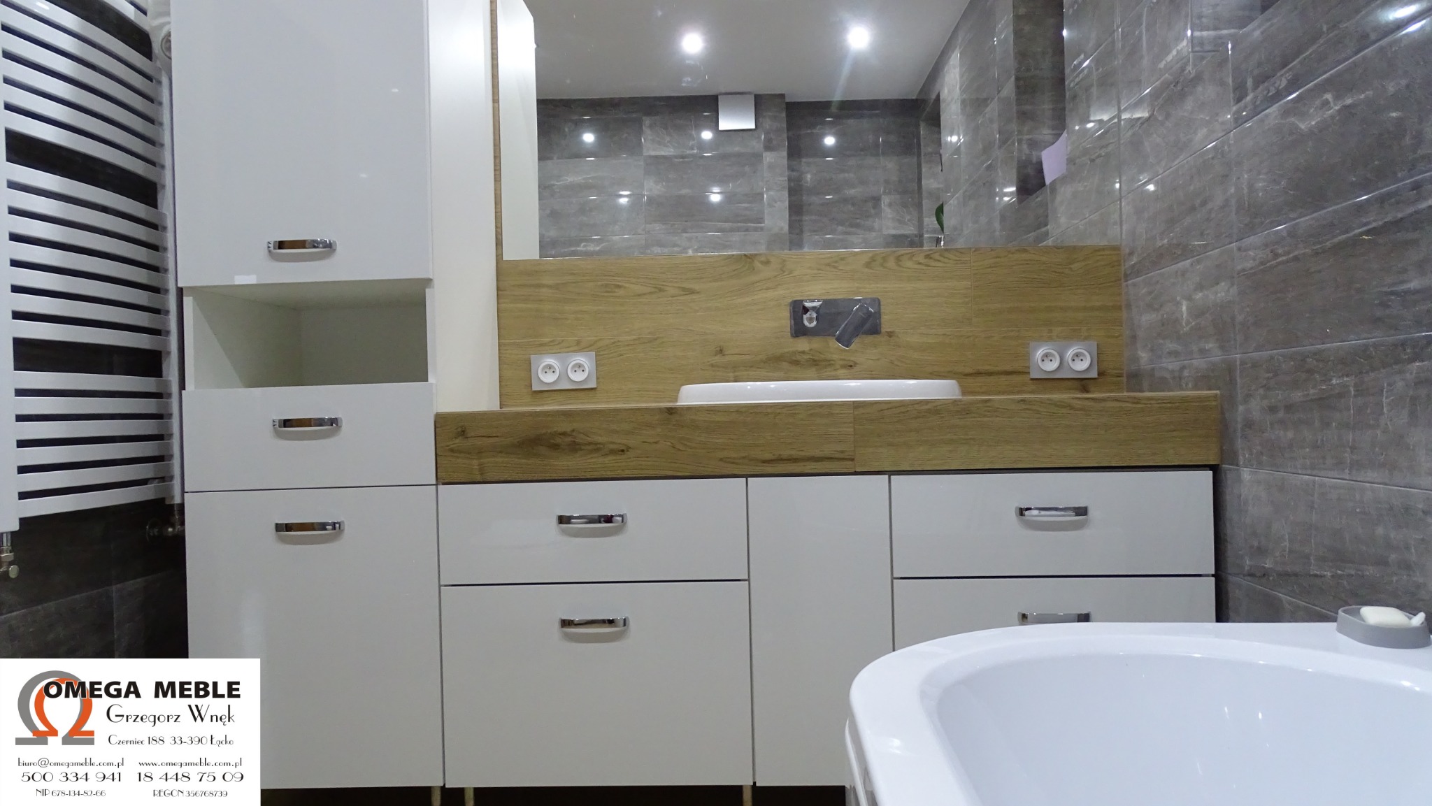 Nowoczesna łazienka z białymi szafkami z szufladami i drewnianym blatem z umywalką wpuszczaną, szare płytki na ścianach, grzejnik drabinkowy po lewej stronie oraz fragment wanny widoczny w prawym...