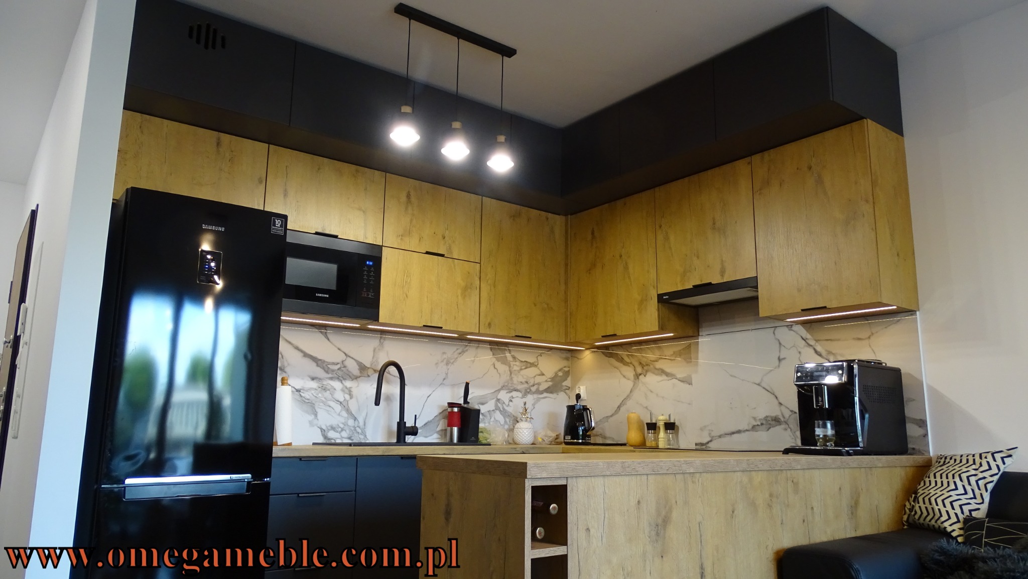 Nowoczesna kuchnia z drewnopodobnymi frontami szafek, czarnymi elementami i marmurowym blatem, oświetlona trzema wiszącymi lampami.
