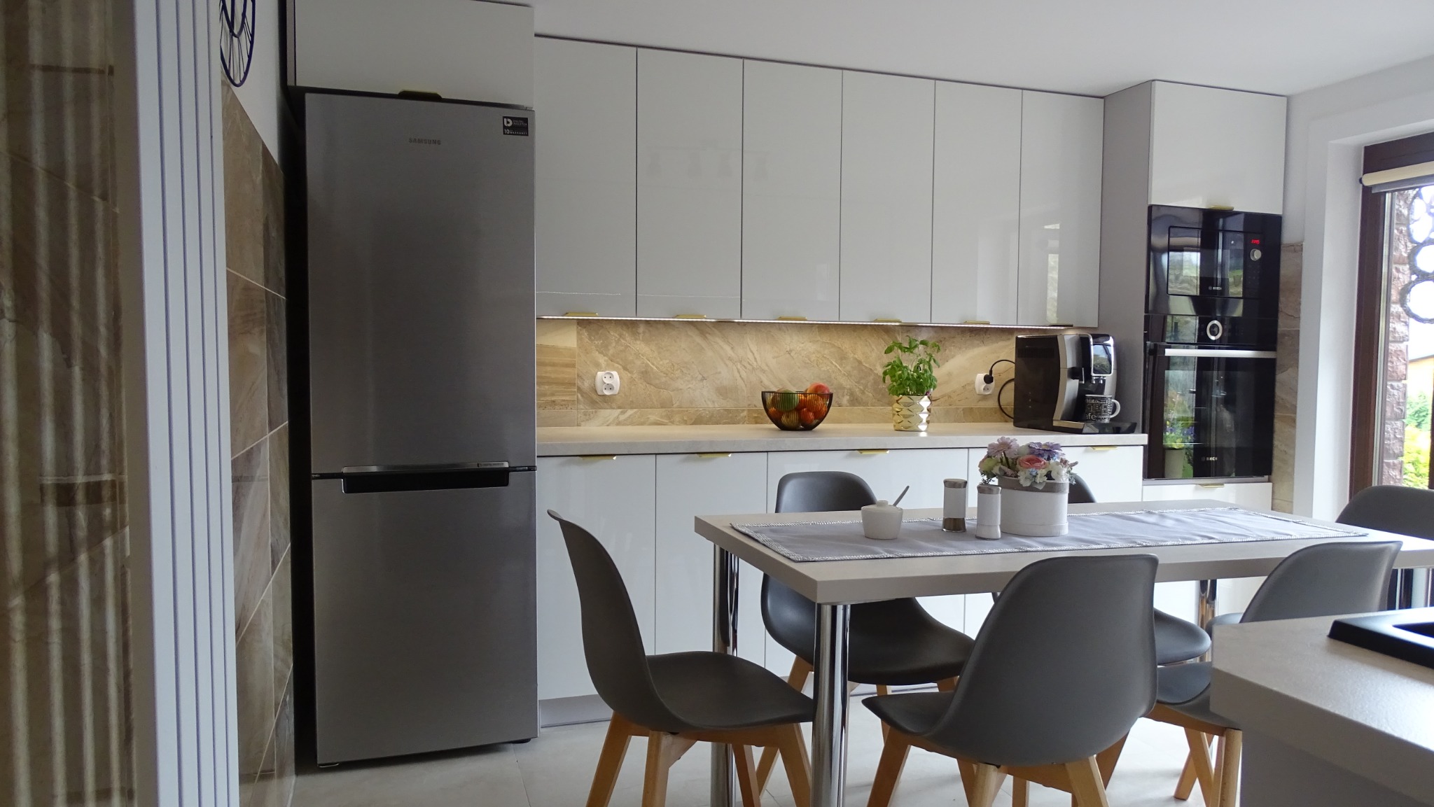Nowoczesna kuchnia z białymi, błyszczącymi szafkami, marmurowym blatem i srebrną lodówką Samsung. Na stole kwiaty w ceramicznej doniczce i szare krzesła.